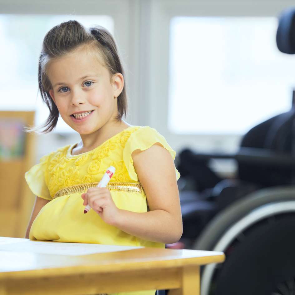 Girl in yellow dress with marker in hand standing at a table in a classroom smiling at camera, wheelchair in the background