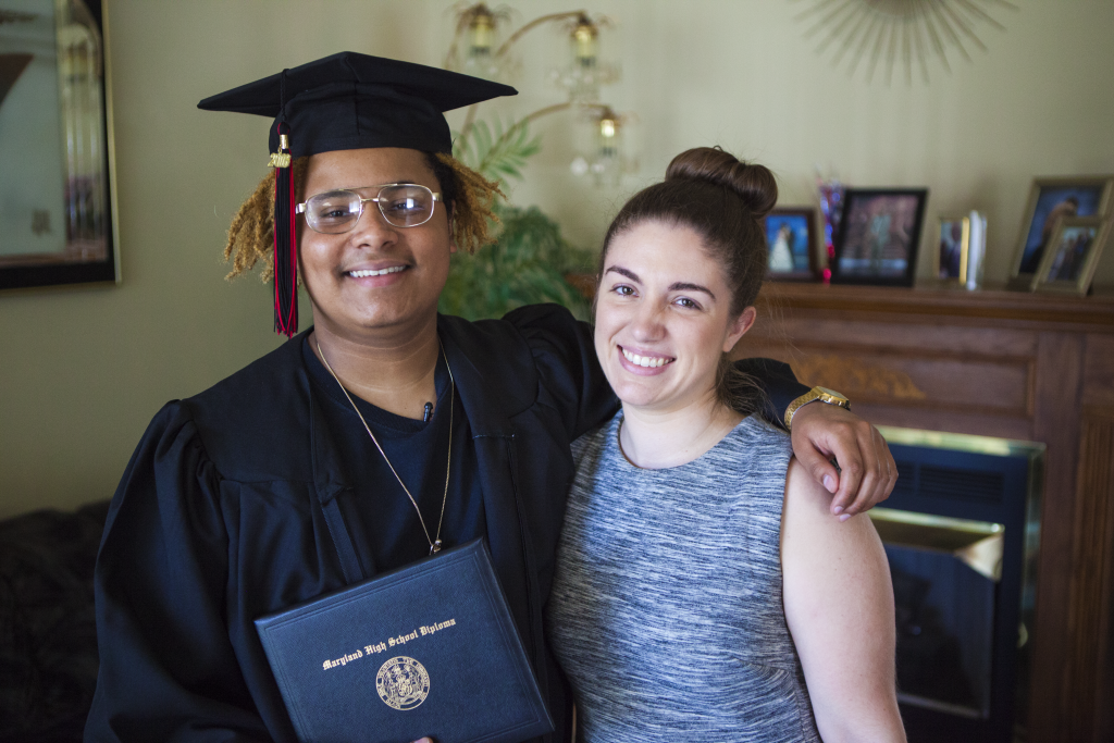 Darius wears his graduation gown and holds his high school diploma. He has his arm around DRM Attorney Amanda White
