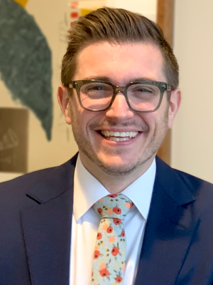 Photo of Anthony May, an attorney at Brown, Goldstein & Levy, LLP. He is a young white man with dark grey glasses, navy blue suit, white shirt, and floral tie. He has short brown hair and facial hair.