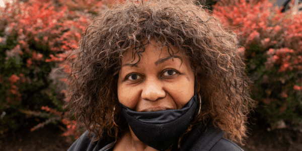 Close up photo of Cookie Lockley, a Black woman with wavy hair. She is wearing a black hoodie, black face mask and surrounded by a red bush.