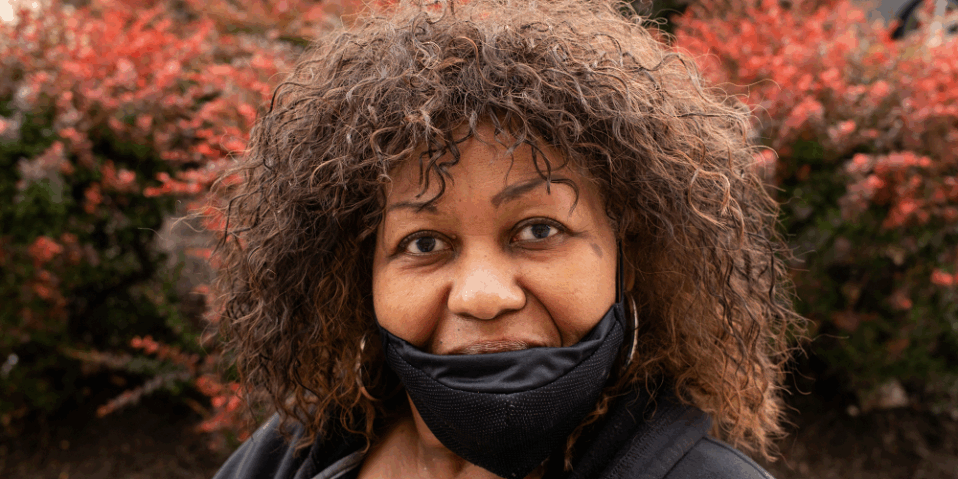 Close up photo of Cookie Lockley, a Black woman with wavy hair. She is wearing a black hoodie, black face mask and surrounded by a red bush.
