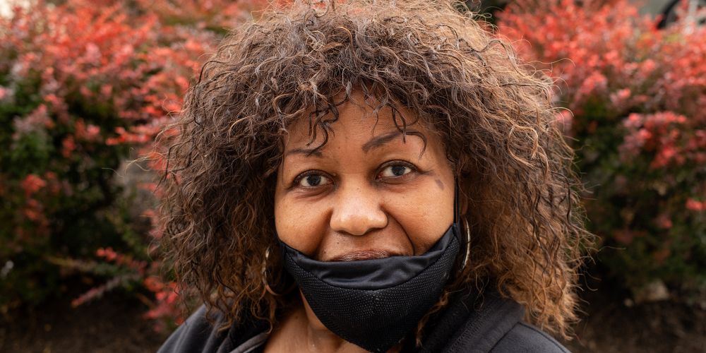 Close up photo of Cookie Lockley, a Black woman with wavy hair. She is wearing a black hoodie, black face mask and surrounded by a red bush.