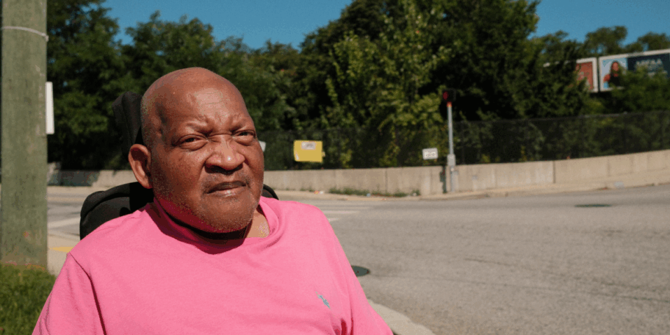 Photo of Floyd Hartley, a Black man with a bald head, using an electric wheelchair and wearing a pink shirt. He is posing near an intersection near his home in NE Baltimore City