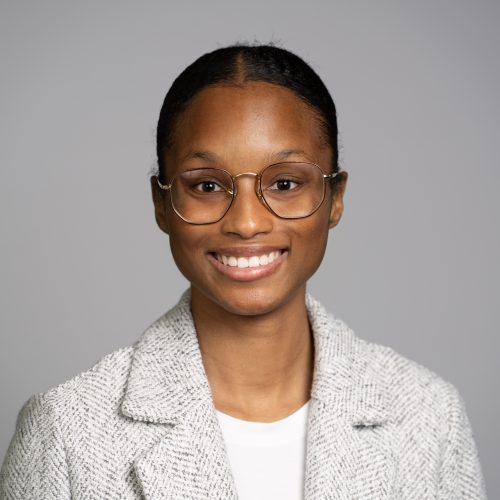 Grace Newton is a young Black woman with her hair tied back. She smiles brightly and wears thin rimmed glasses, a white blouse, and a light gray blazer.