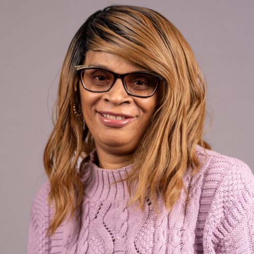 Jacqueline Phillips is a Black woman with long golden brown hair. She smiles wearing a pink sweater and gold and black rimmed, tinted glasses.