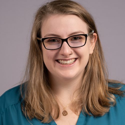 Megan Jones is a young White woman with long light brown hair and thin nose rings. She smiles brightly wearing black rimmed glasses, and a teal vneck blouse.