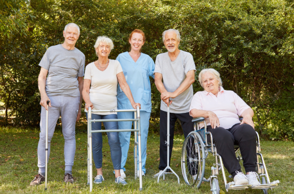 4 older adults outside smiling (one in a wheelchair, one with a walker, and two with canes) and a worker