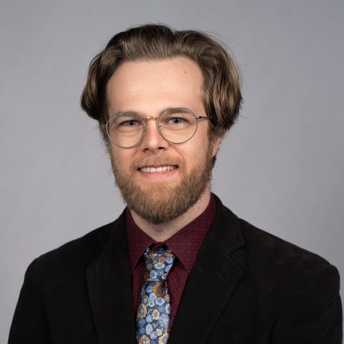 Phil McGuire is a young White man with mid length, straight brown hair and a full beard and mustache. He smiles wearing thin, silver rimmed glasses, a black corduroy blazer, red patterned collared shirt and floral tie.