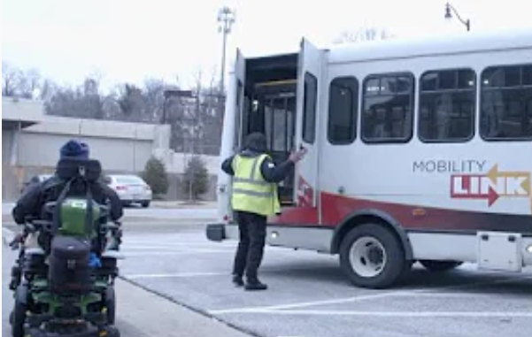 Motorized wheelchair user approaching a Mobility Link van with an attendant opening the van doors.