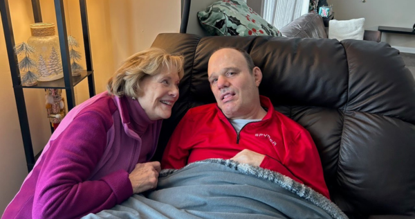 Photo of Linda and her son Troy. Linda is a older White woman with short blonde hair wearing a purple sweater and squatting down next to Troy and smiling at him. Troy is an older White man sitting in a sofa chair. He is wearing a red sweater and sitting under a blue blanket.