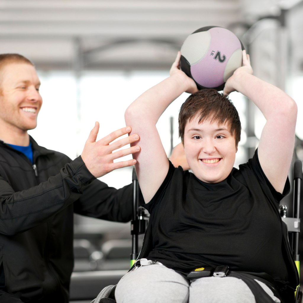 Woman at the gym in wheelchair lifting exercise ball behind her head with a male trainer close by