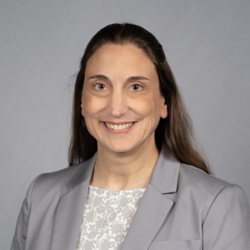 Tam Kelley is a White woman with long brown hair. She smiles wearing a light grey blazer and grey and cream patterned blouse.