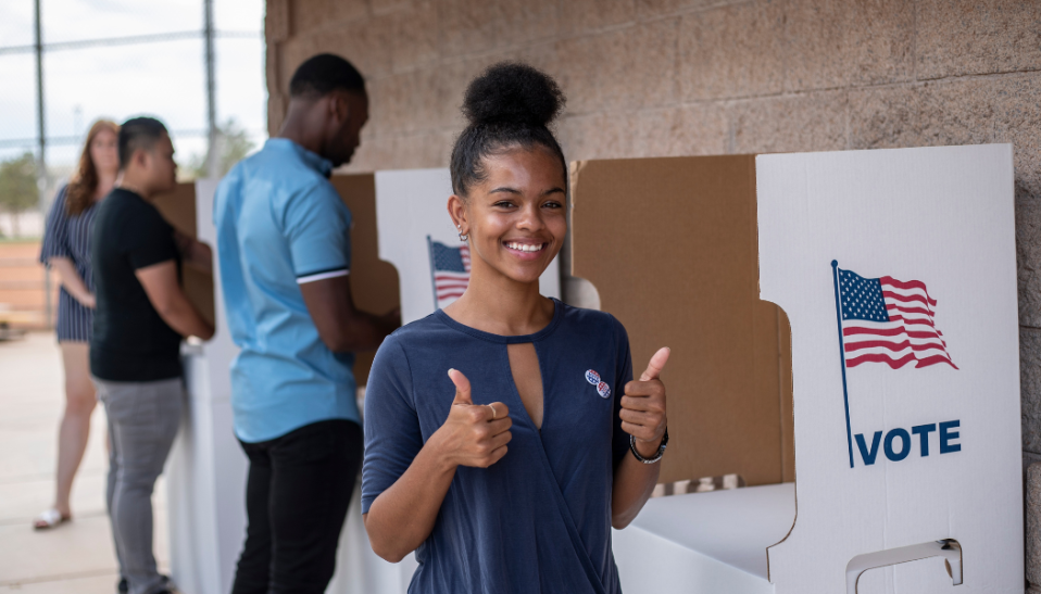 Black woman standing in front of voting booth with thumbs up and smiling at the camera