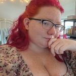 Photo of Sage Viohl a young white woman with black rimmed glasses, red hair, and brown patterned blouse