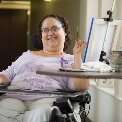 Woman in wheelchair smiling holding a paint brush sitting in front of a canvas