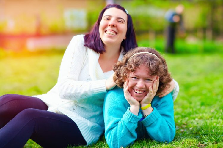 Two women lounging on a green lawn. They are both smiling, and one has her hands on the shoulders of the other.