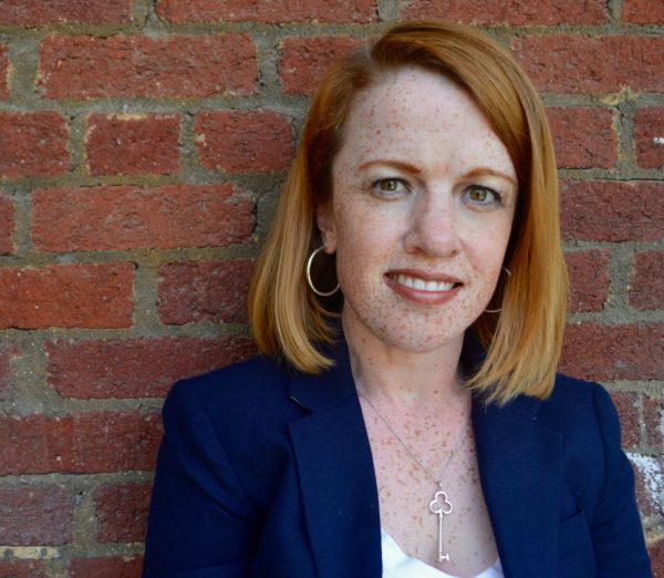 Photo of Rebecca Cokley. White woman with red hair wearing a navy blazer and white tank top, standing in front of a brick wall.